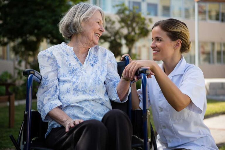 An elderly resident sits in a comfortable room with a loved one holding their hand and a caregiver nearby.
